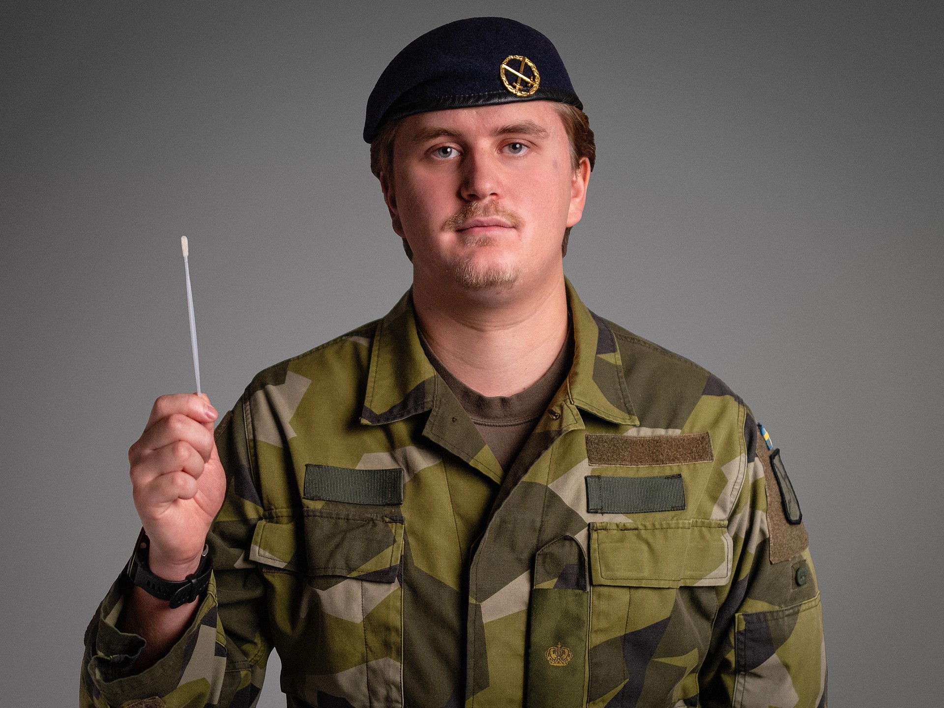 En soldat i kamouflageuniform och mörk basker håller i en medicinsk provtagningspinne.