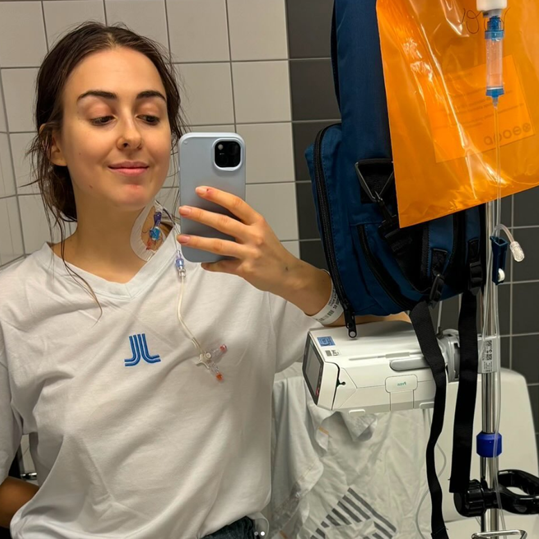 En ung kvinna med en intravenös slang fäst vid sitt bröst och ett sjukhusarmband ler svagt medan hon tar en selfie i spegeln, med en droppställning och pump i bakgrunden.