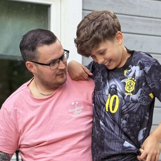 En man i rosa skjorta tittar på en leende pojke i mönstrad skjorta, som har armen runt mannens axel.