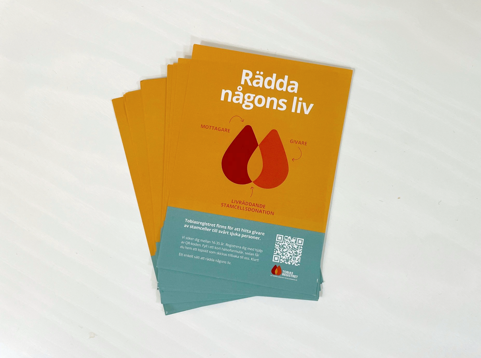 A stack of orange and teal brochures promoting stem cell donation, featuring interconnected blood-like drops and the Swedish text "Rädda någons liv" (Save someone's life).