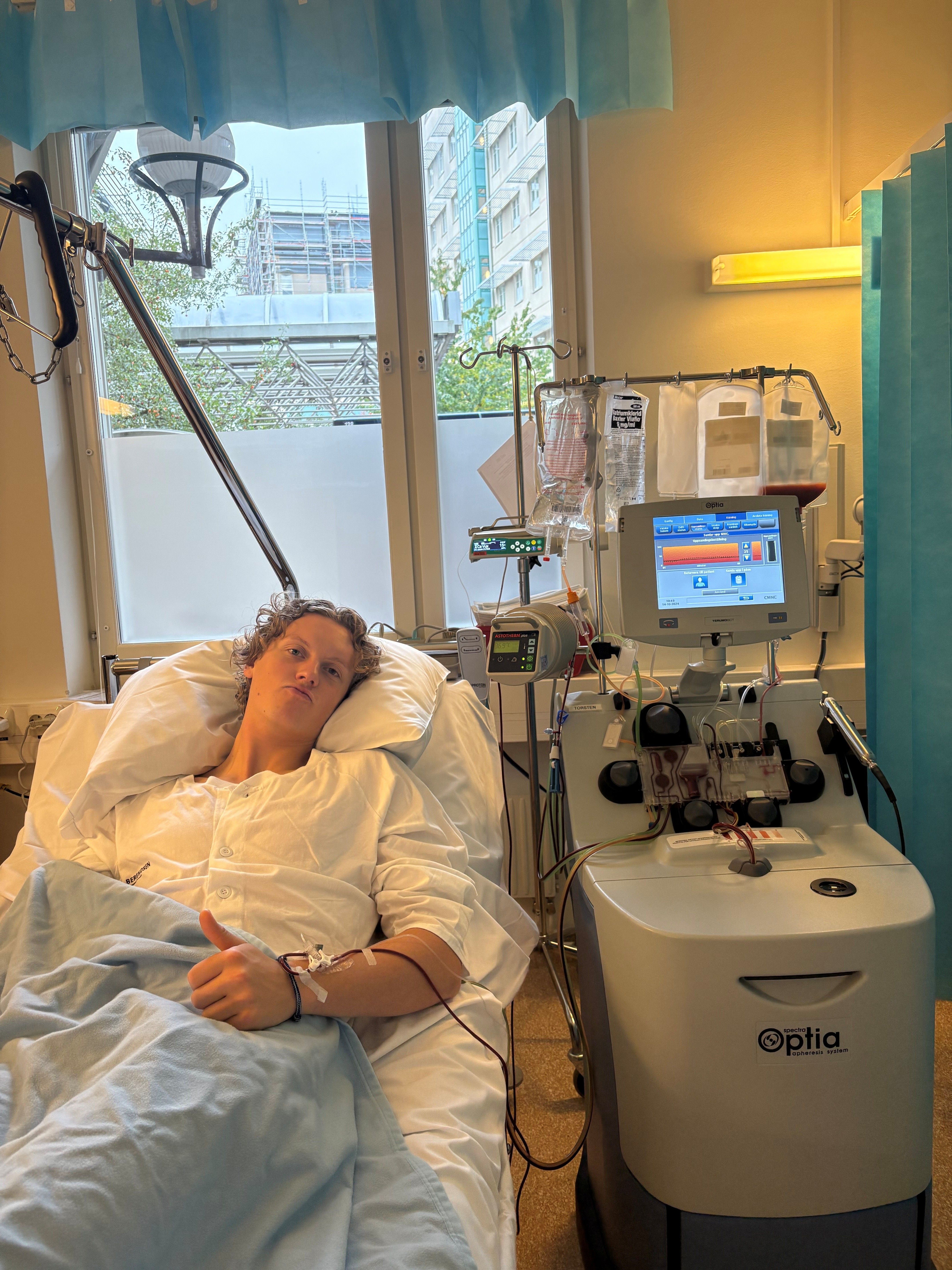 En patient i en sjukhussäng ger tummen upp medan hen är kopplad till en Optia-aferesmaskin.