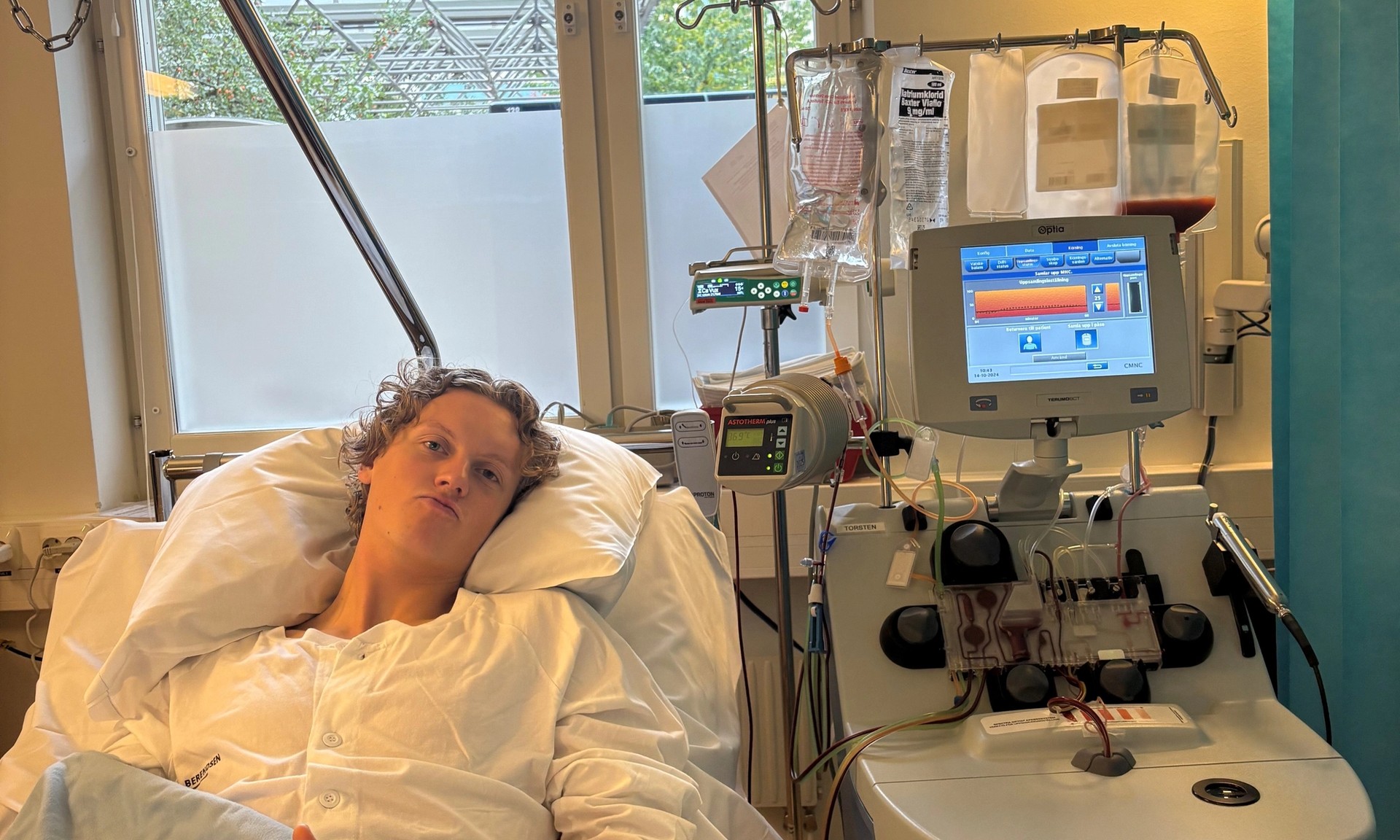 En patient i en sjukhussäng ger tummen upp medan hen är kopplad till en Optia-aferesmaskin.