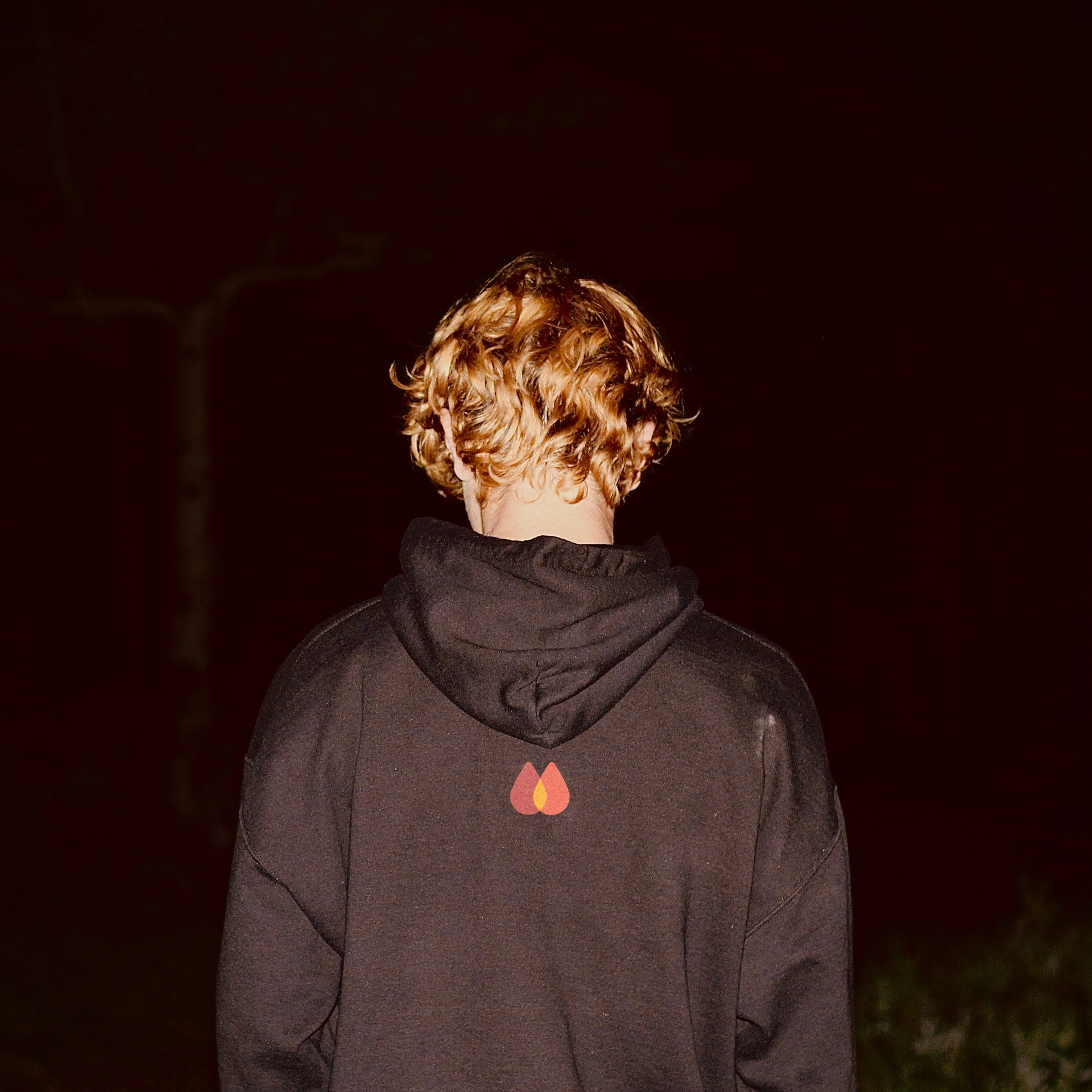 Back of a person with curly reddish-blonde hair, wearing a dark hoodie with a red and yellow flame-like logo, against a dark background.