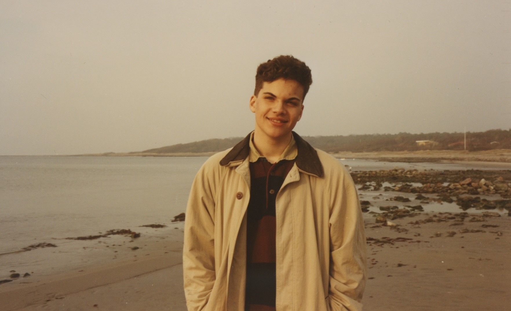 Young man in a beige jacket smiling on a cloudy beach.