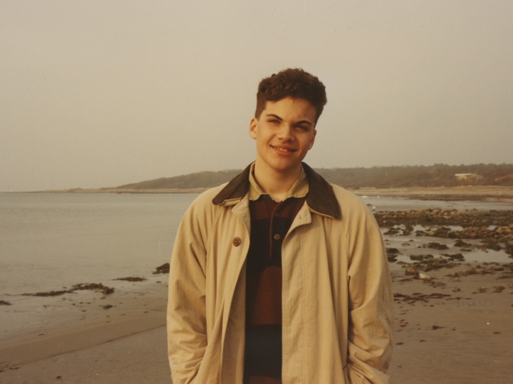 Young man in a beige jacket smiling on a cloudy beach.