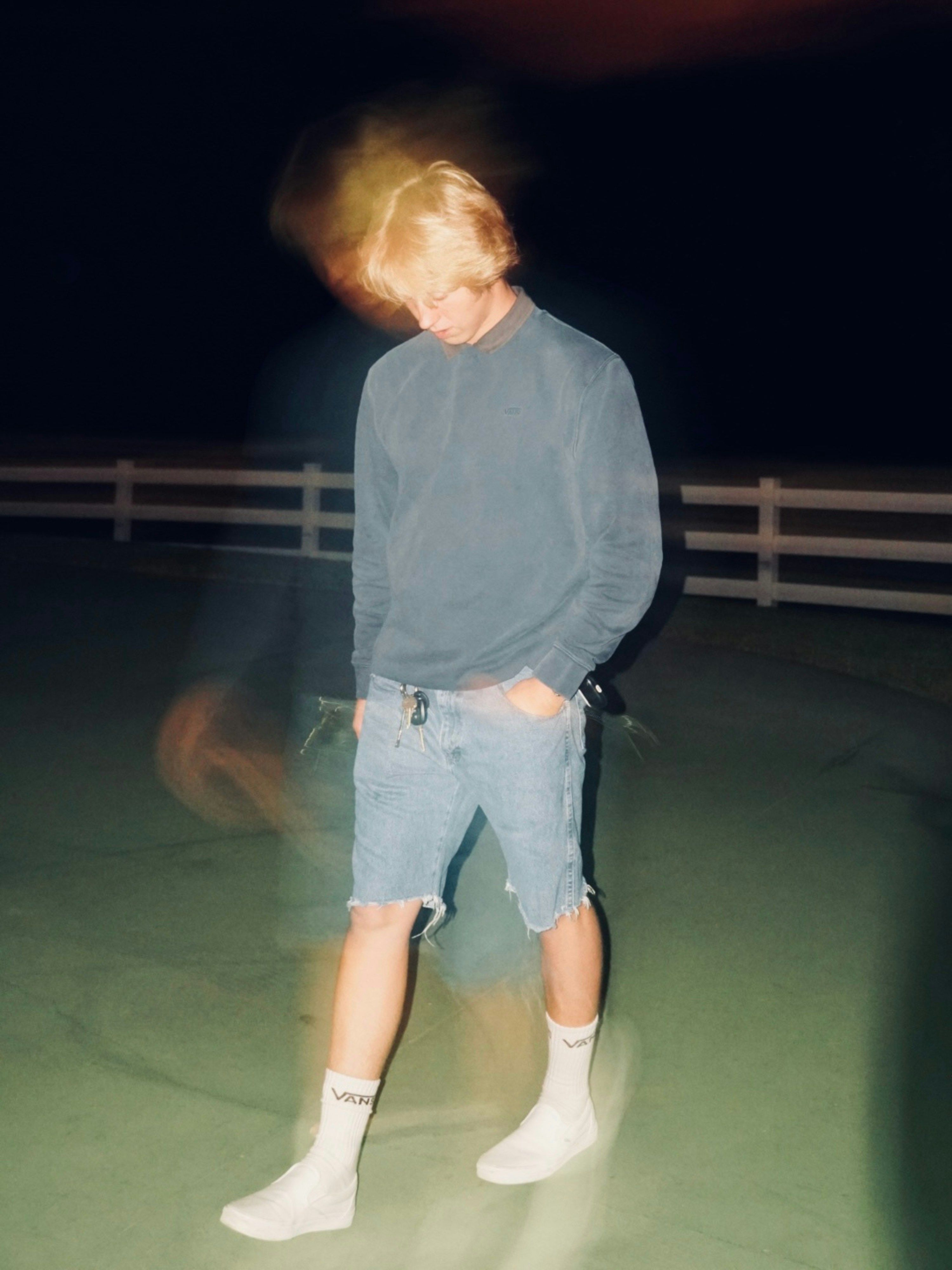 A blond-haired person in a blue sweatshirt, cutoff denim shorts, and white Vans socks stands looking down at night, with significant motion blur.