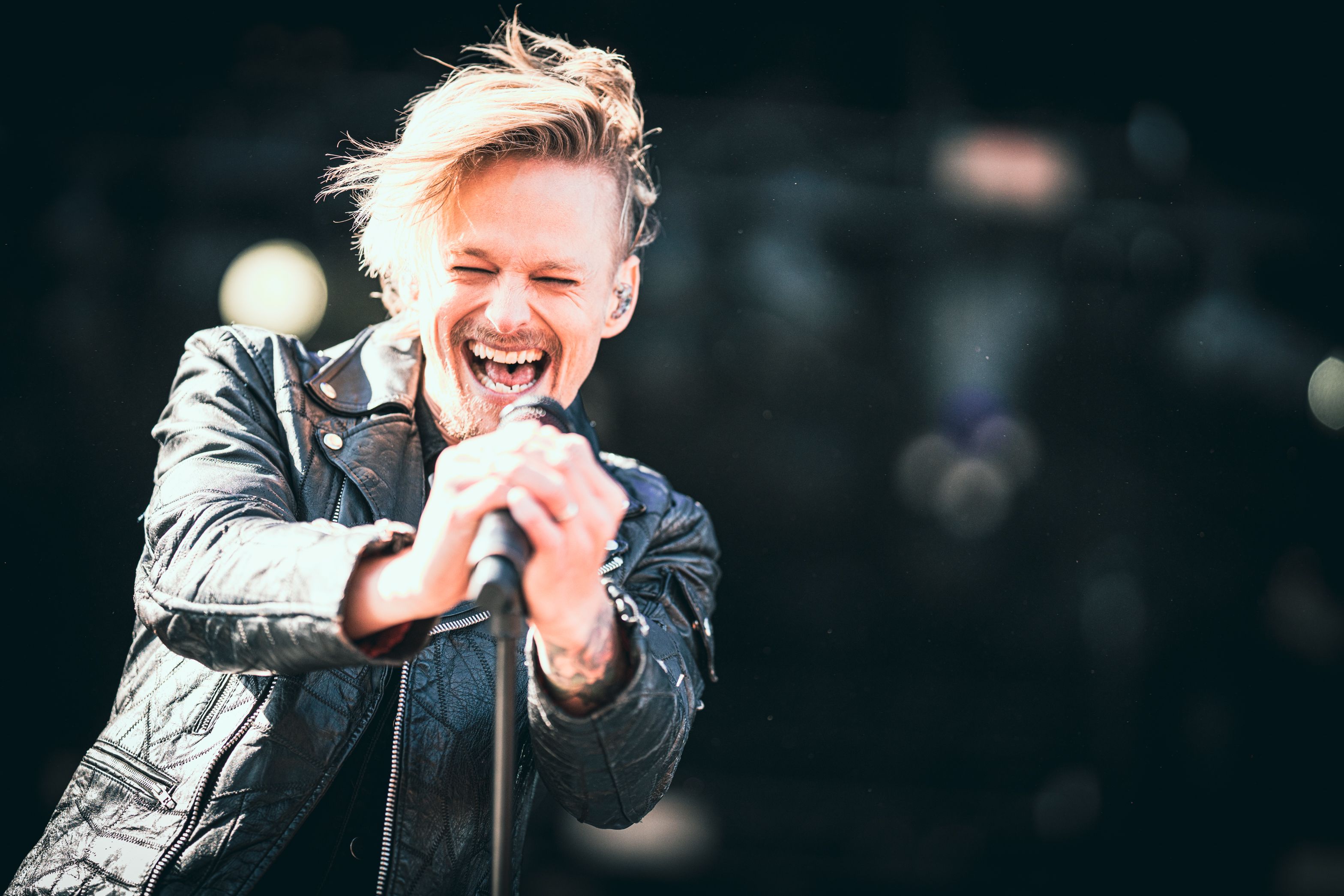 A male singer with blonde hair and a leather jacket joyfully sings into a microphone, eyes closed, mouth wide open.