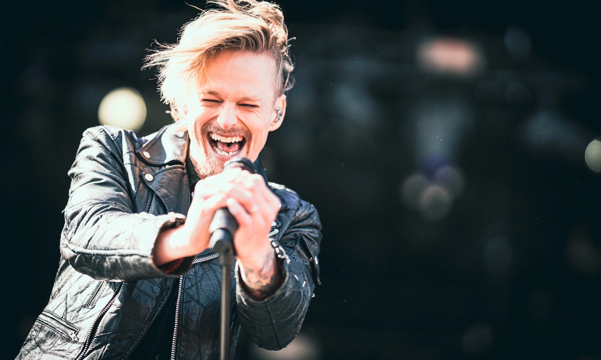 A male singer with blonde hair and a leather jacket joyfully sings into a microphone, eyes closed, mouth wide open.