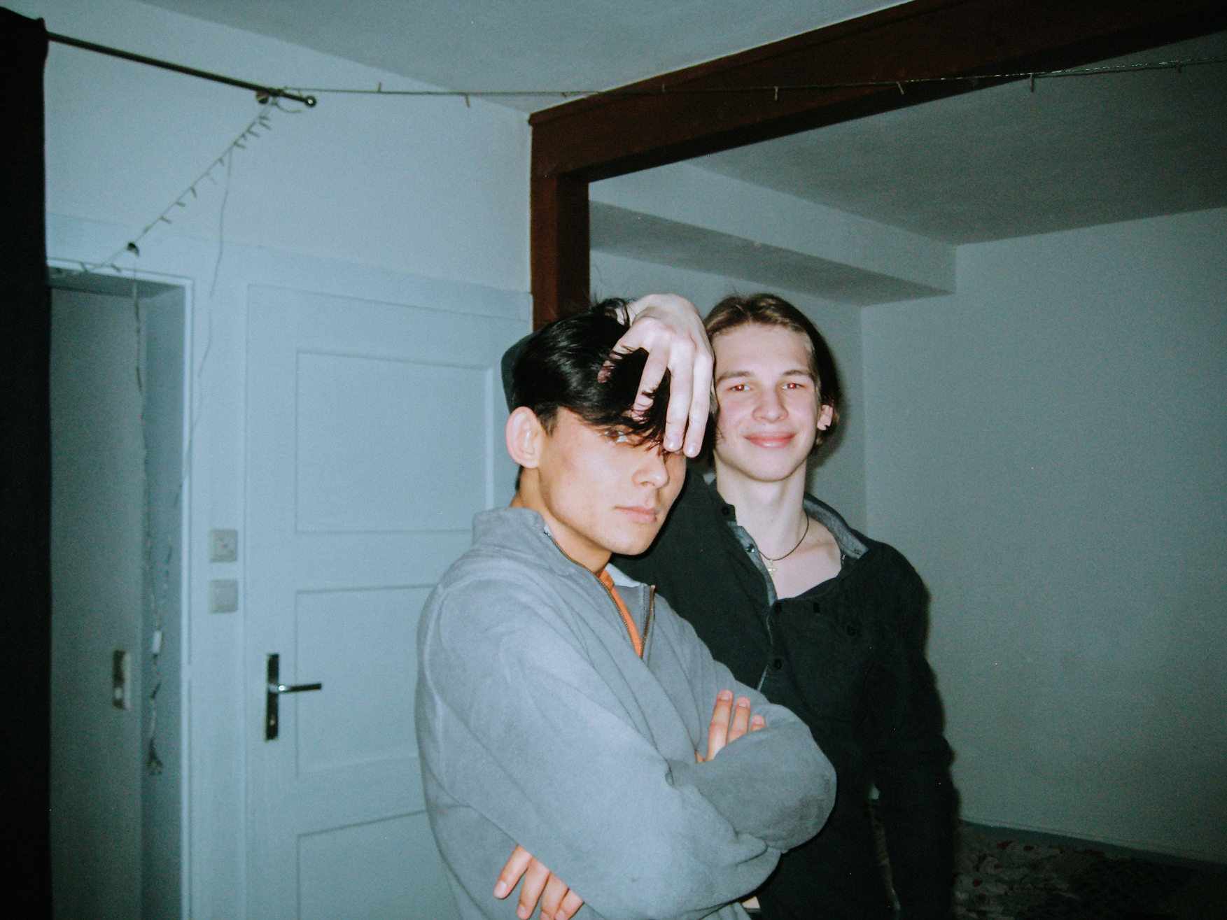 Två unga män poserar inomhus; den ena ler med en hand på den andres huvud, som har korsade armar.