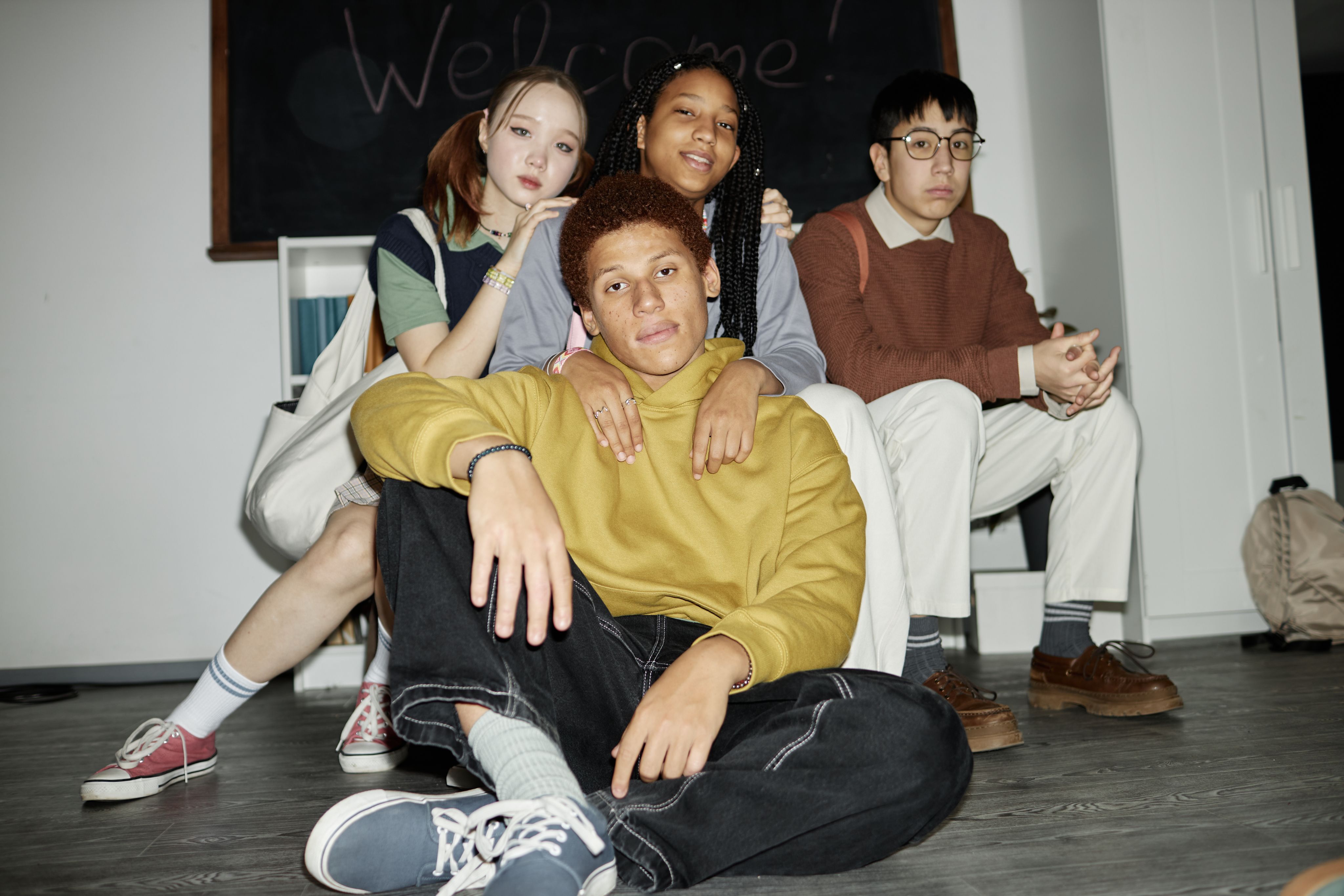 Four diverse young people pose indoors, with one sitting on the floor and three behind him, against a "Welcome!" blackboard.