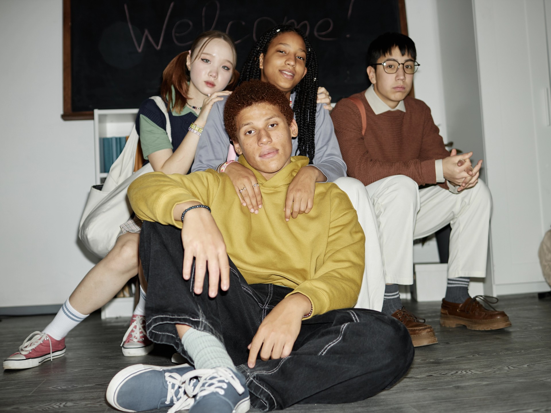 Four diverse young people pose indoors, with one sitting on the floor and three behind him, against a "Welcome!" blackboard.
