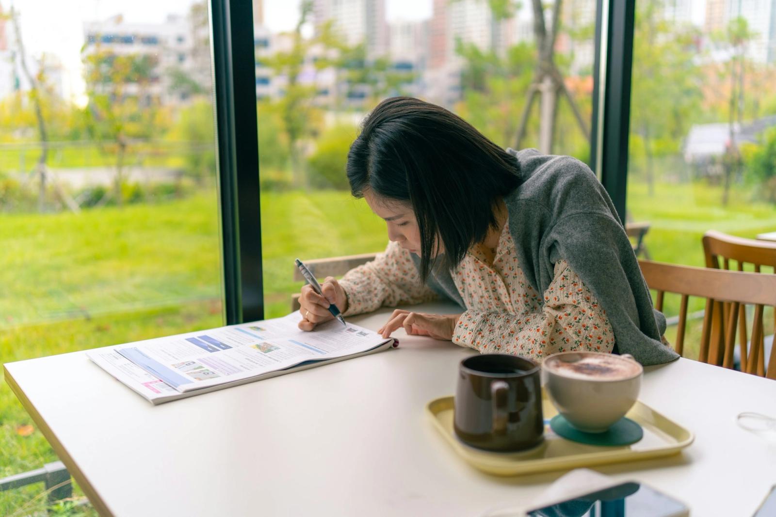 An adult woman writing in a notebook at a sunlit table by a garden window.