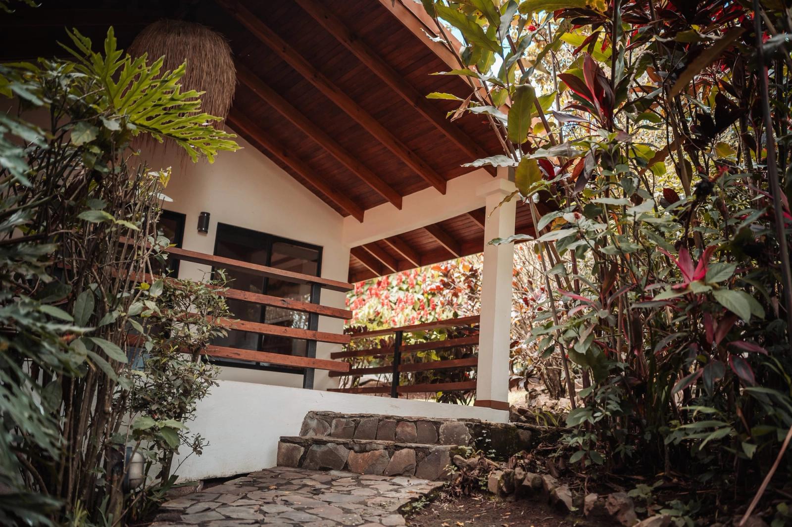 Un pabellón blanco con techo de madera y escalones de piedra, enmarcado por plantas tropicales en los jardines de Atenas.