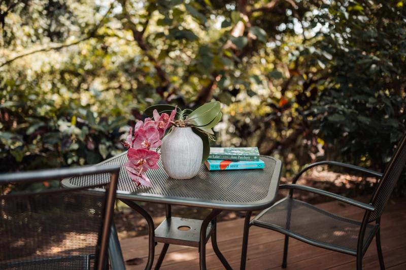 A metal garden table with an orchid and books on a wooden deck, surrounded by greenery.