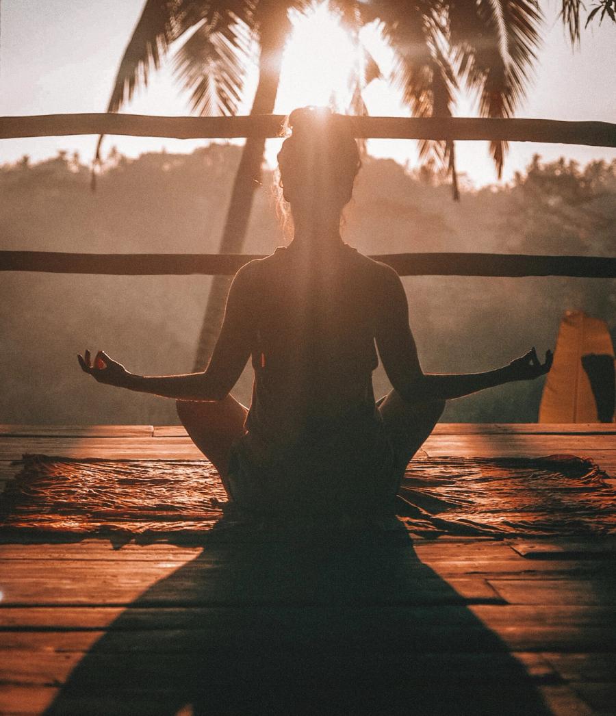 A person sits in meditation on a wooden deck, silhouetted against the late-day sun, with palms overhead and the green hills beyond.