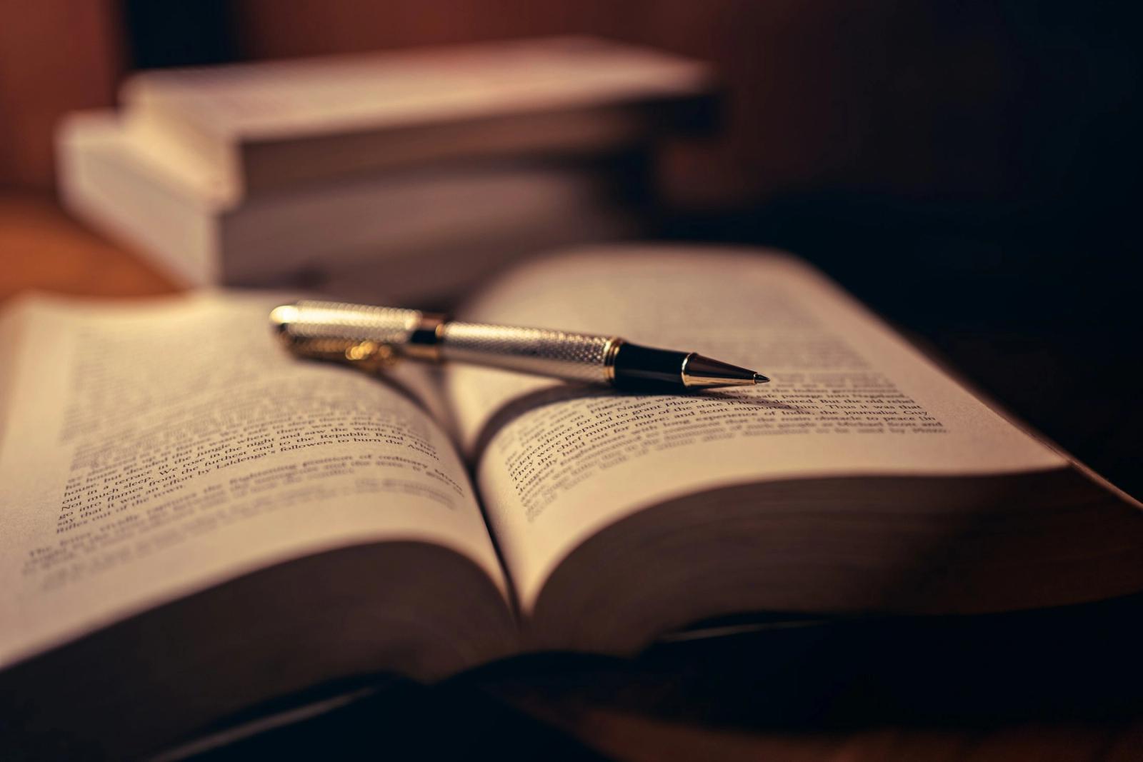 An open book with a pen resting on it in warm lamplight, a small stack of books behind.