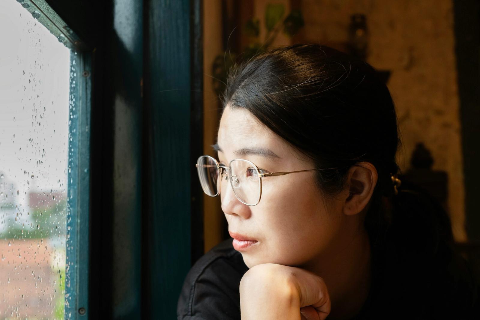 A woman rests her chin on her hand, gazing out a rain-flecked window in warm light.