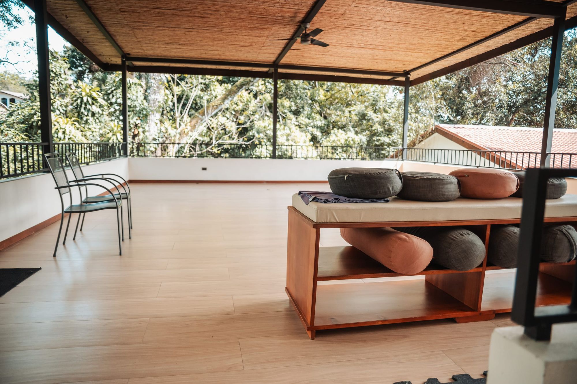 Cushions on a wooden bench in the open-air yoga hall, looking out to treetops and a garden chair at the rail.