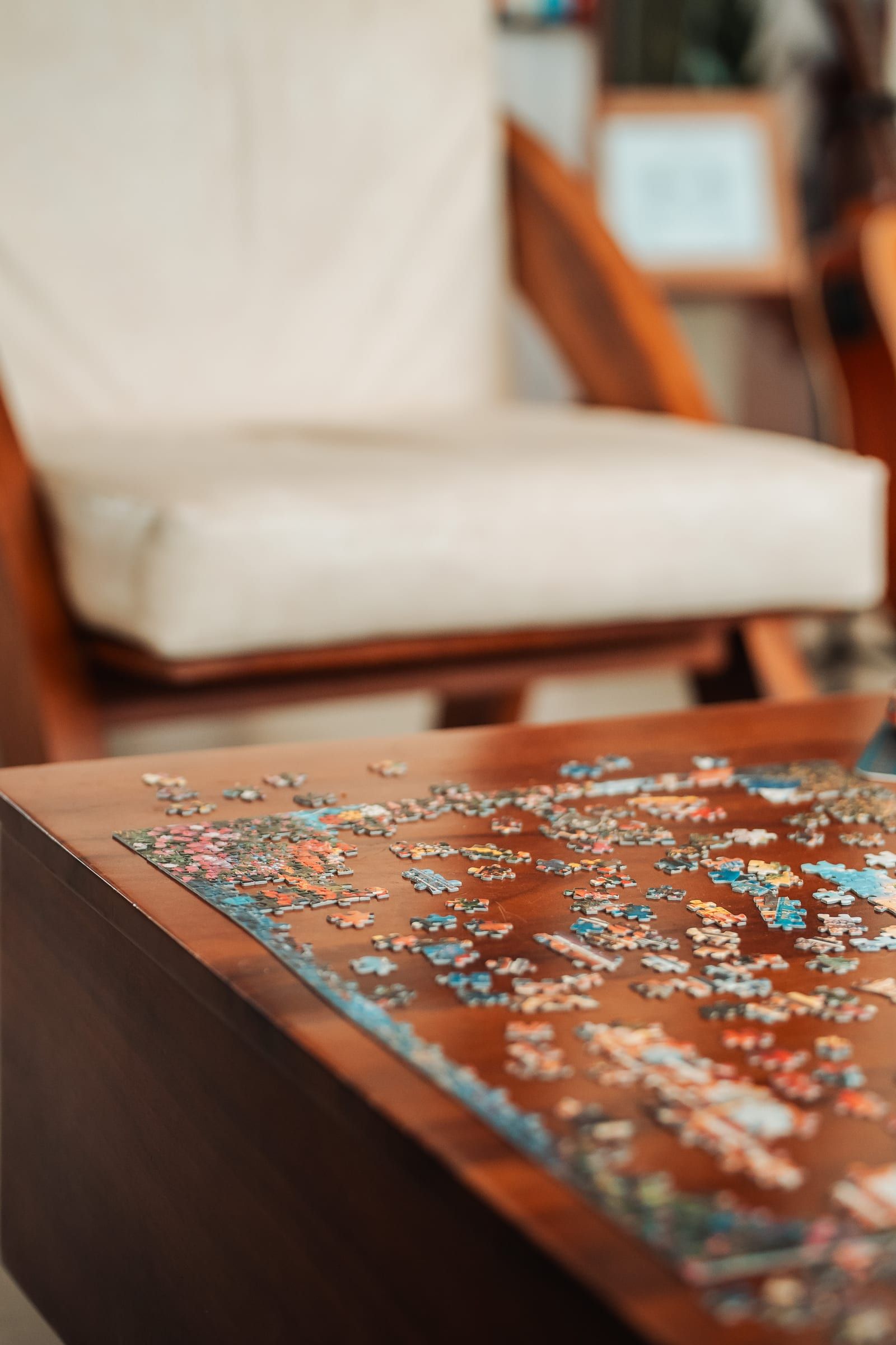 Close-up of jigsaw pieces spread across a wooden table beside a white armchair.