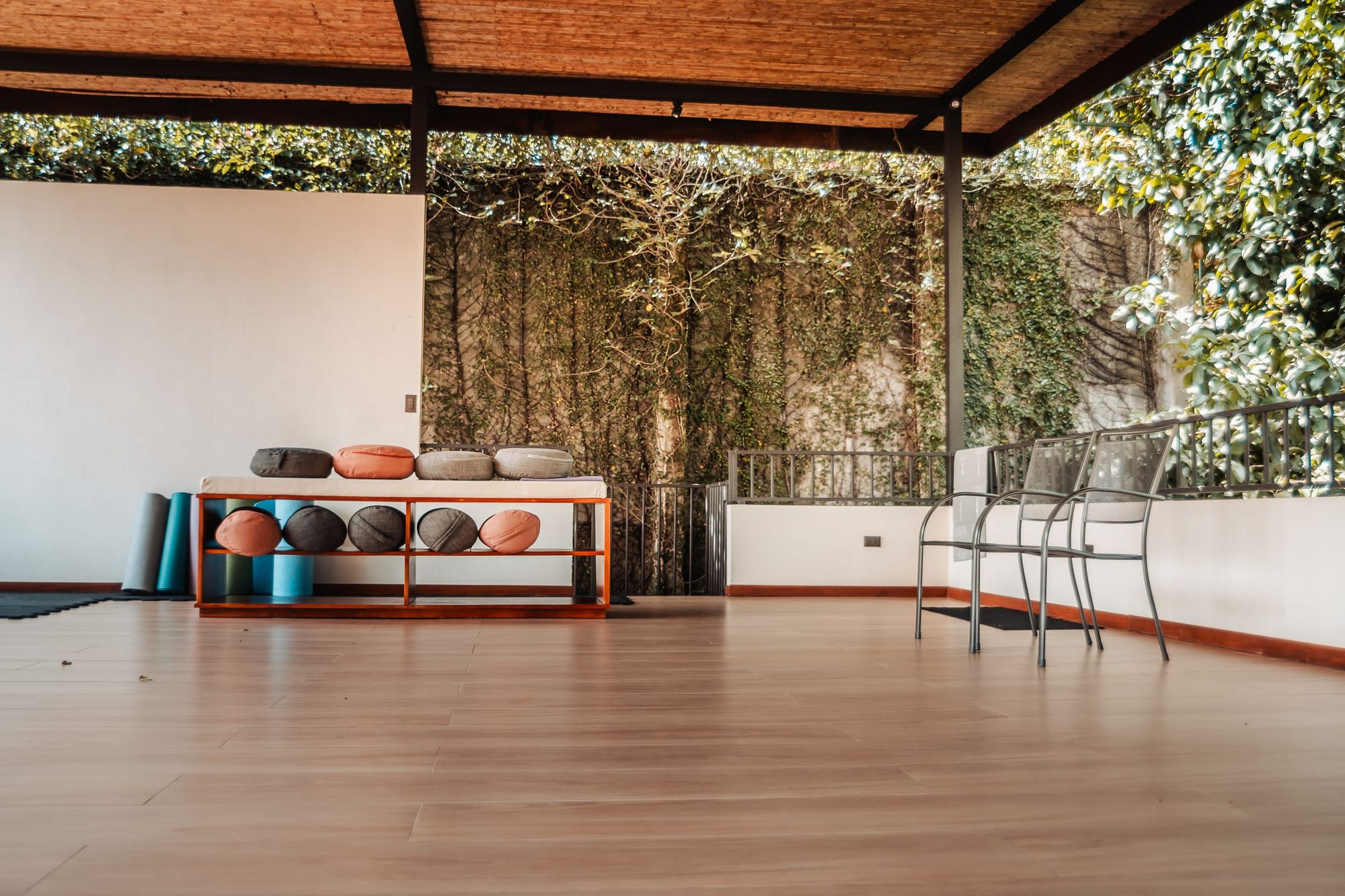 The yoga hall floor with a bench of meditation cushions and mats along a wall covered in climbing plants.