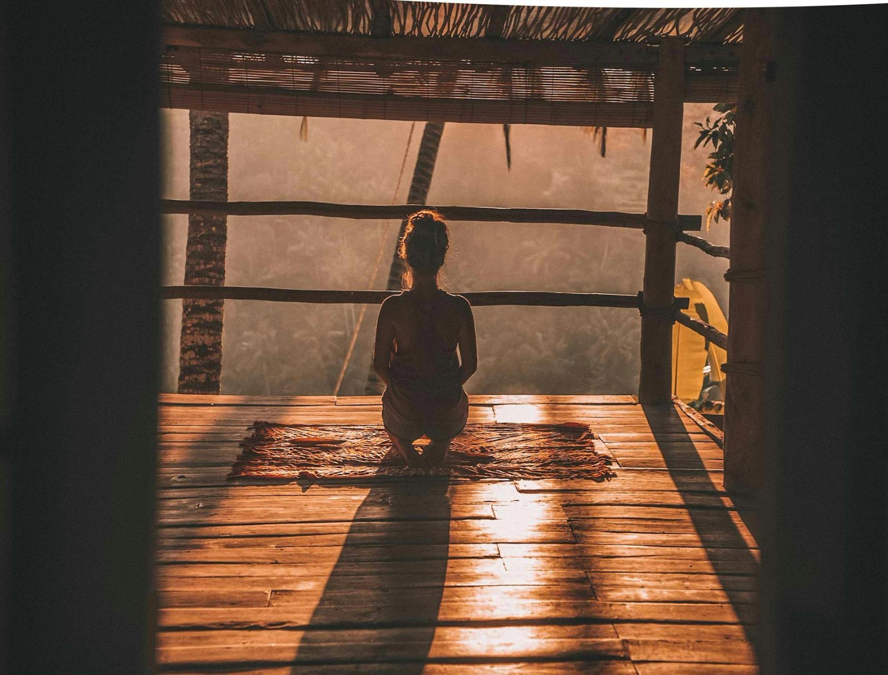 A person sits in quiet meditation, looking out over green treetops in soft morning light.