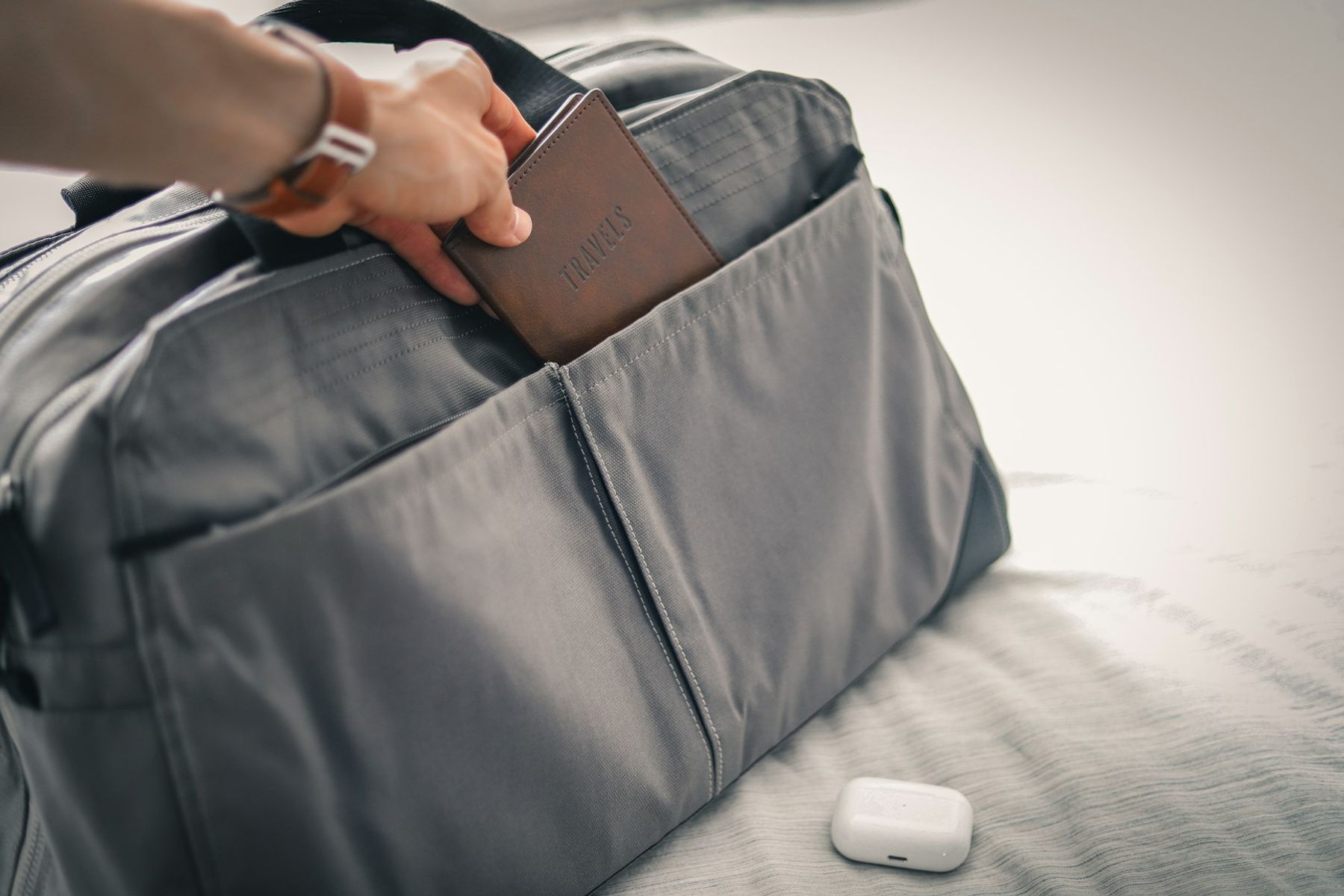 A hand tucking a leather travel document holder into the side pocket of a packed grey weekender bag.