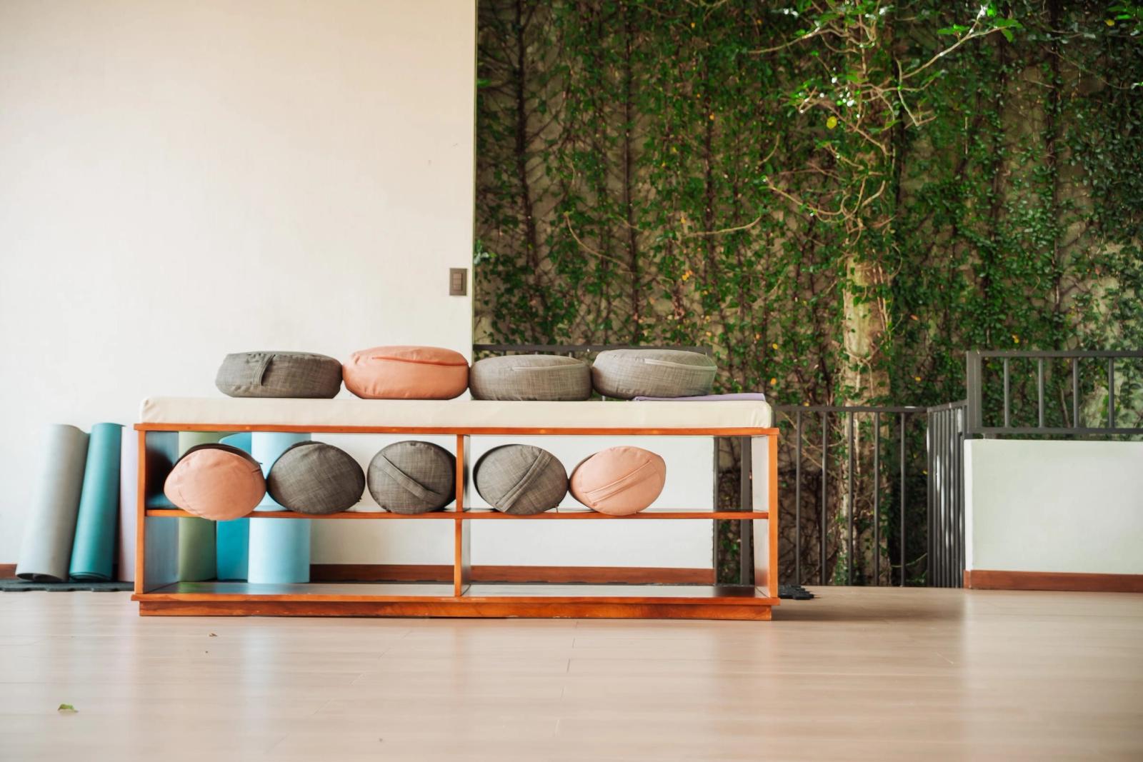 Meditation cushions and rolled yoga mats on a wooden bench in the open-air yoga hall, with a wall of greenery behind.
