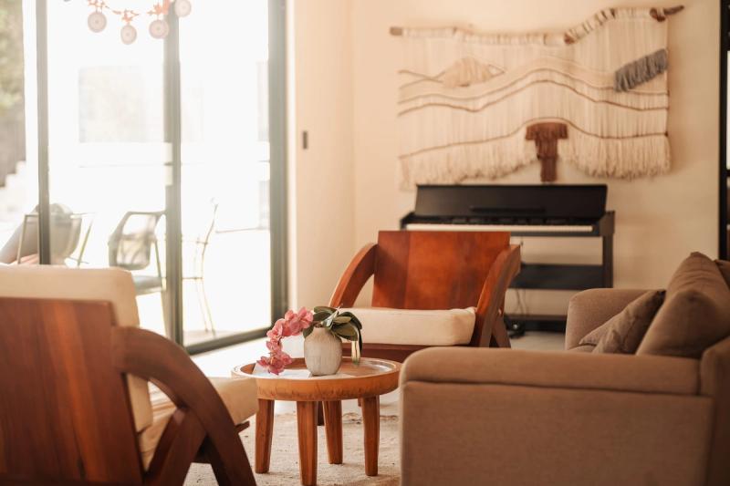 A lounge at Atenas with sofas, wooden armchairs, a round table with flowers and a piano under a wall hanging.