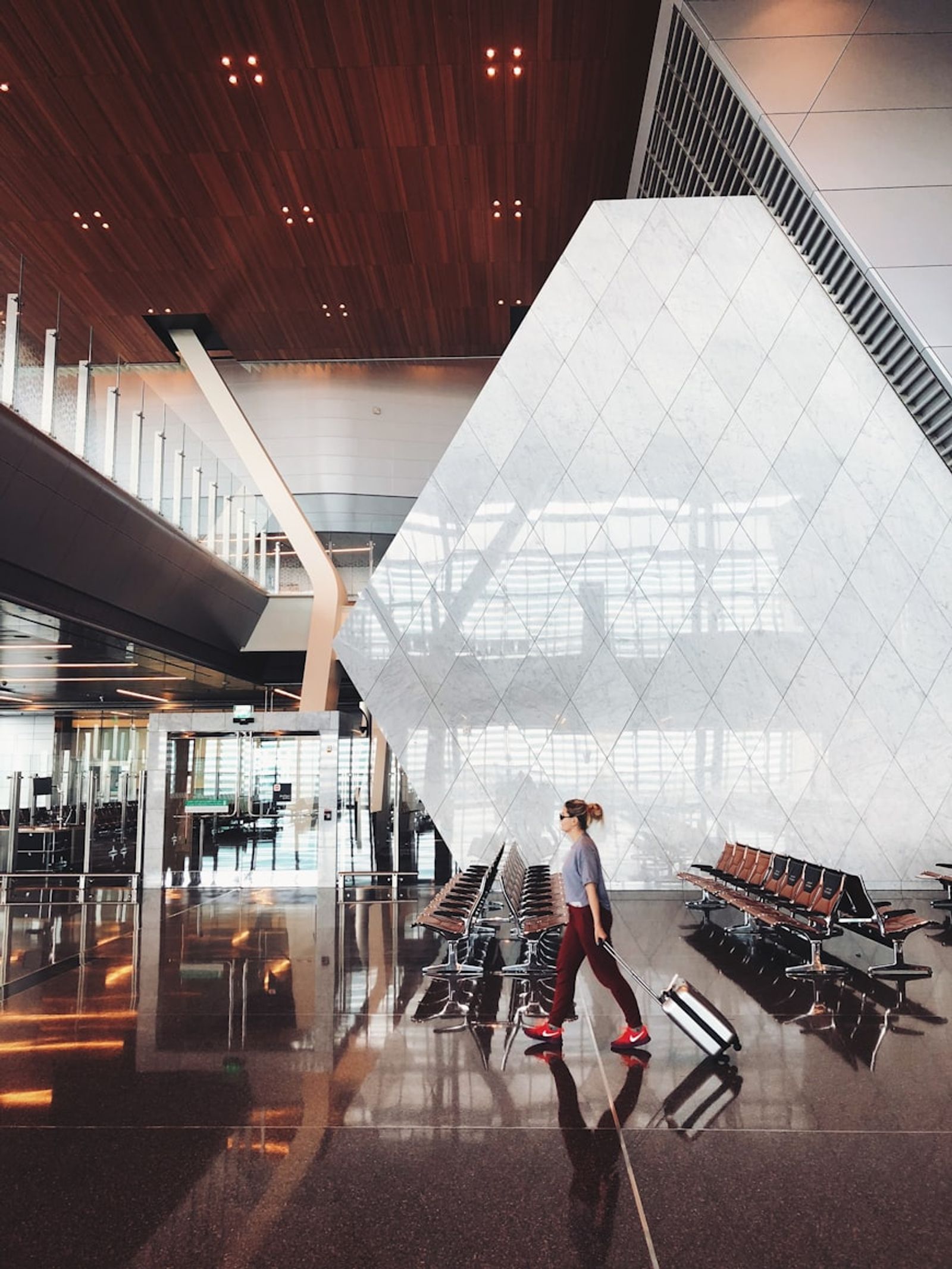 A traveler walking through a bright, modern airport terminal pulling a wheeled suitcase past rows of empty seats.