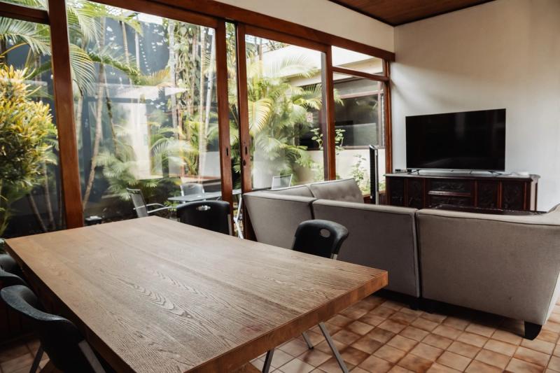 A wooden dining table and a sofa facing a television, beside windows onto the courtyard.