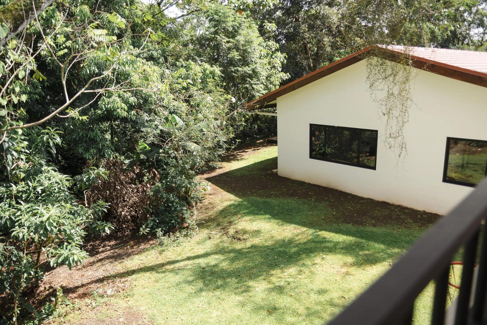 A white house at the edge of a sloping lawn, half hidden by forest, seen from a balcony.