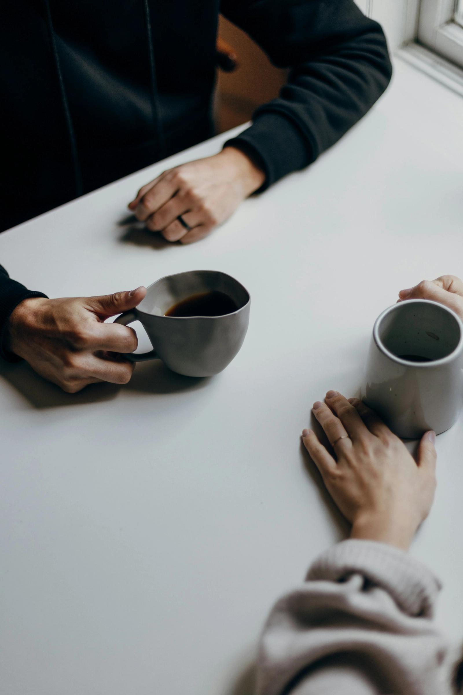 Two people sitting across a table with coffee mugs, hands resting on the surface in quiet conversation.