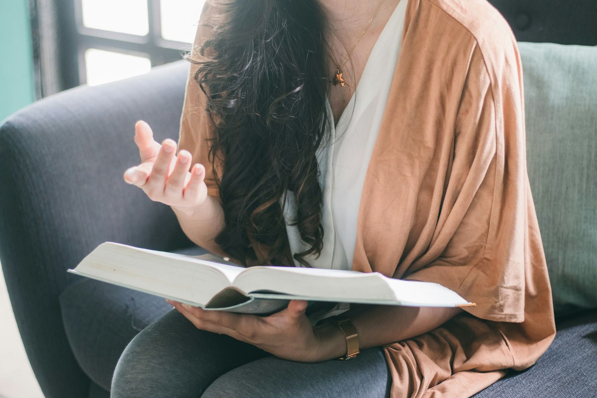 A woman sits on a sofa with an open book in her lap, one hand raised mid-thought in a quiet moment of reflection.