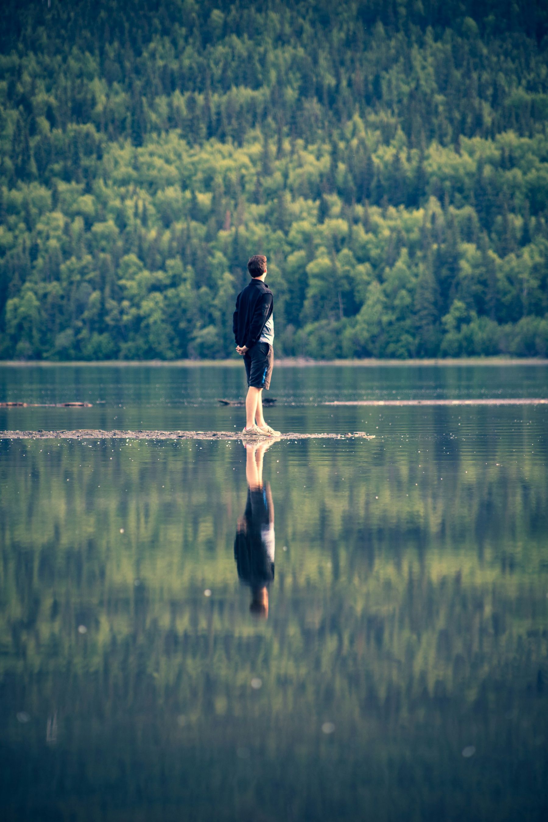 A person stands at the edge of a still lake, reflected in the calm water.