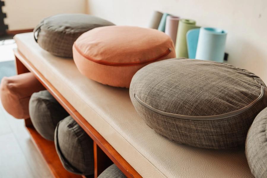 Close-up of round linen meditation cushions stacked on a wooden bench, with yoga mats leaning in the corner.