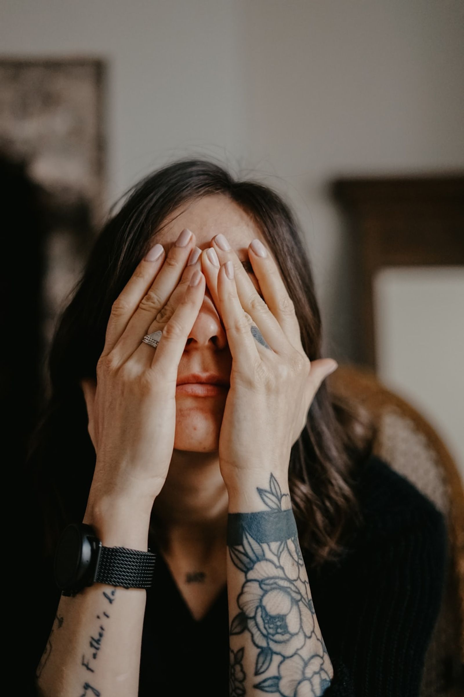 A woman sits in a dimly lit room with both hands pressed over her face, a gesture of overwhelm.
