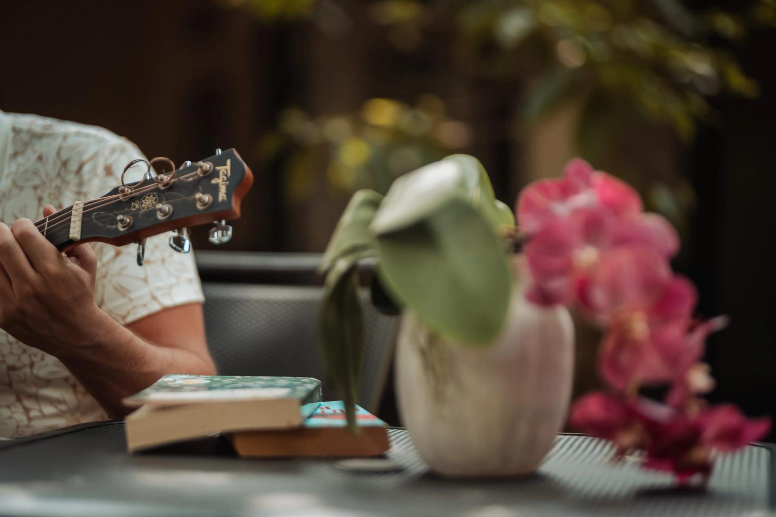 Hands playing a guitar at a garden table, next to an orchid and a pair of books.