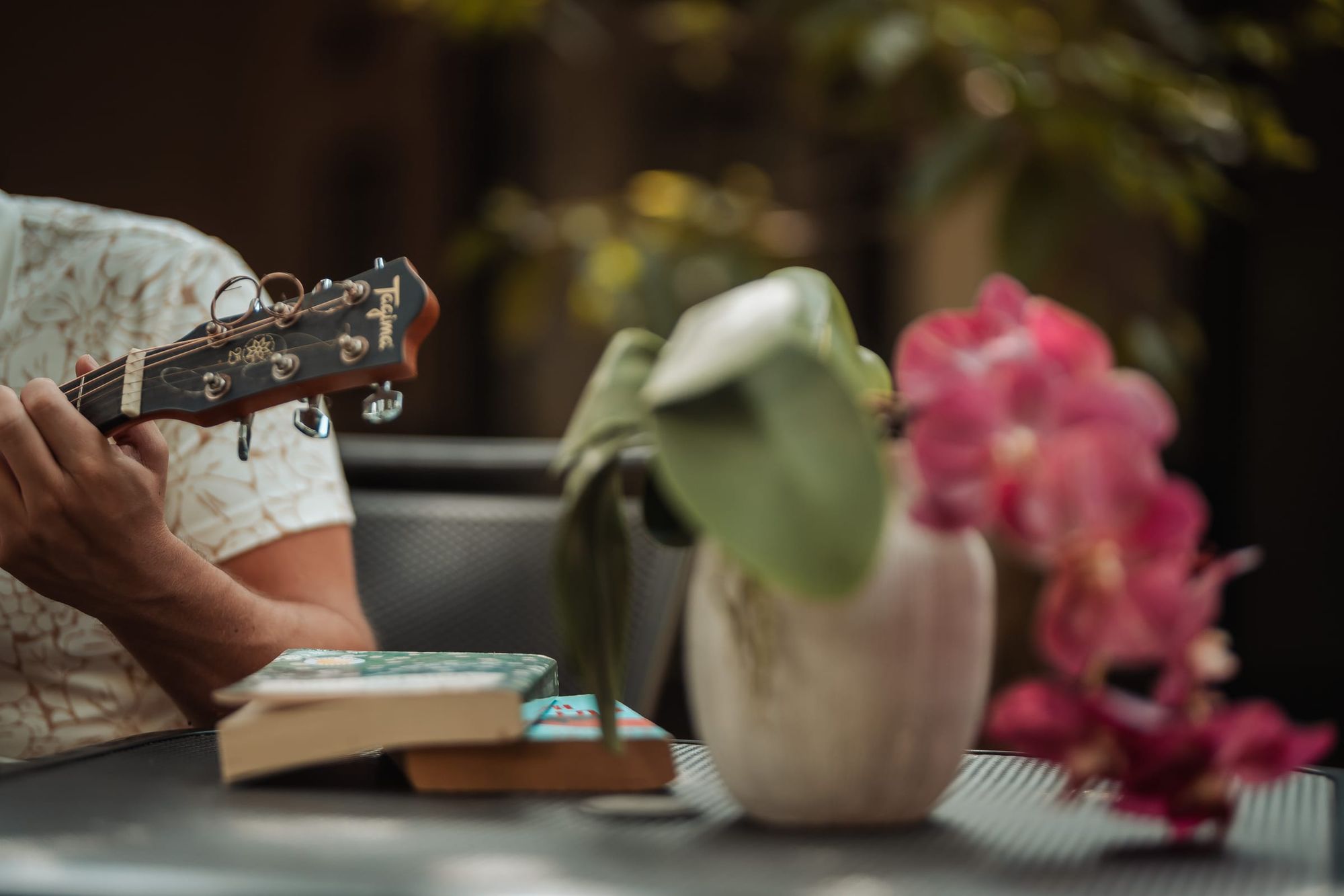 Unas manos tocan la guitarra en una mesa del jardín, junto a una orquídea y un par de libros.