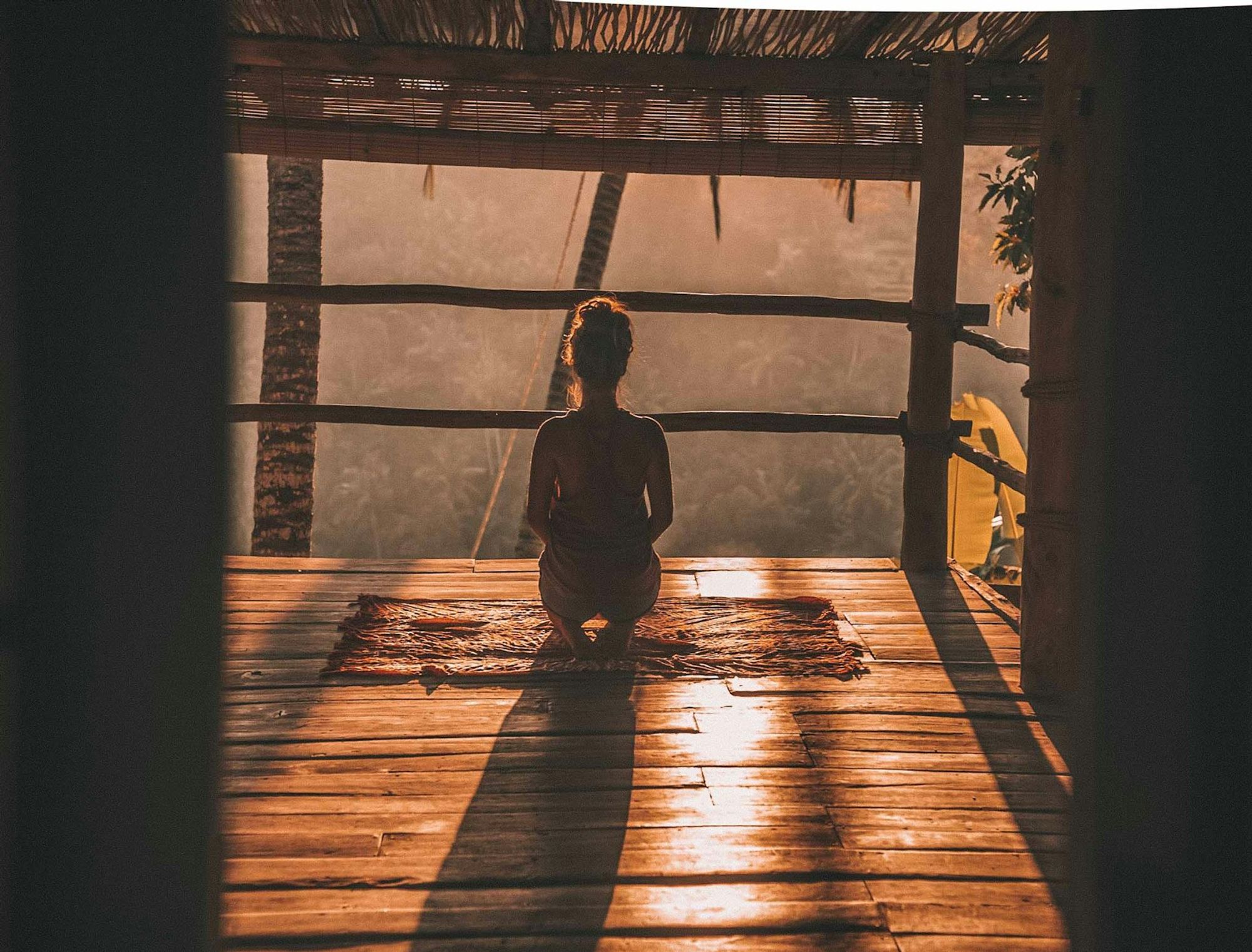 A person kneels in silhouette on a wooden deck at golden hour, meditating, with forest beyond an open-air pavilion.