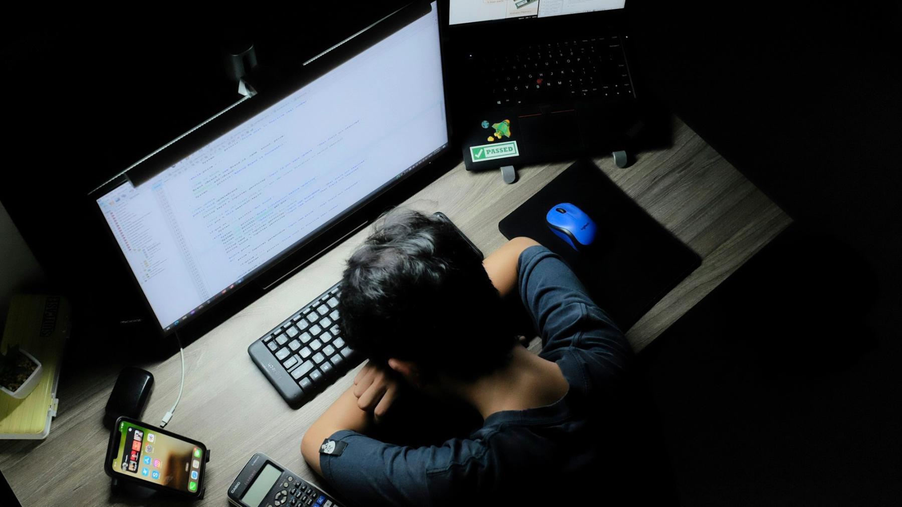 A person resting their head on a desk at night beside glowing computer screens.