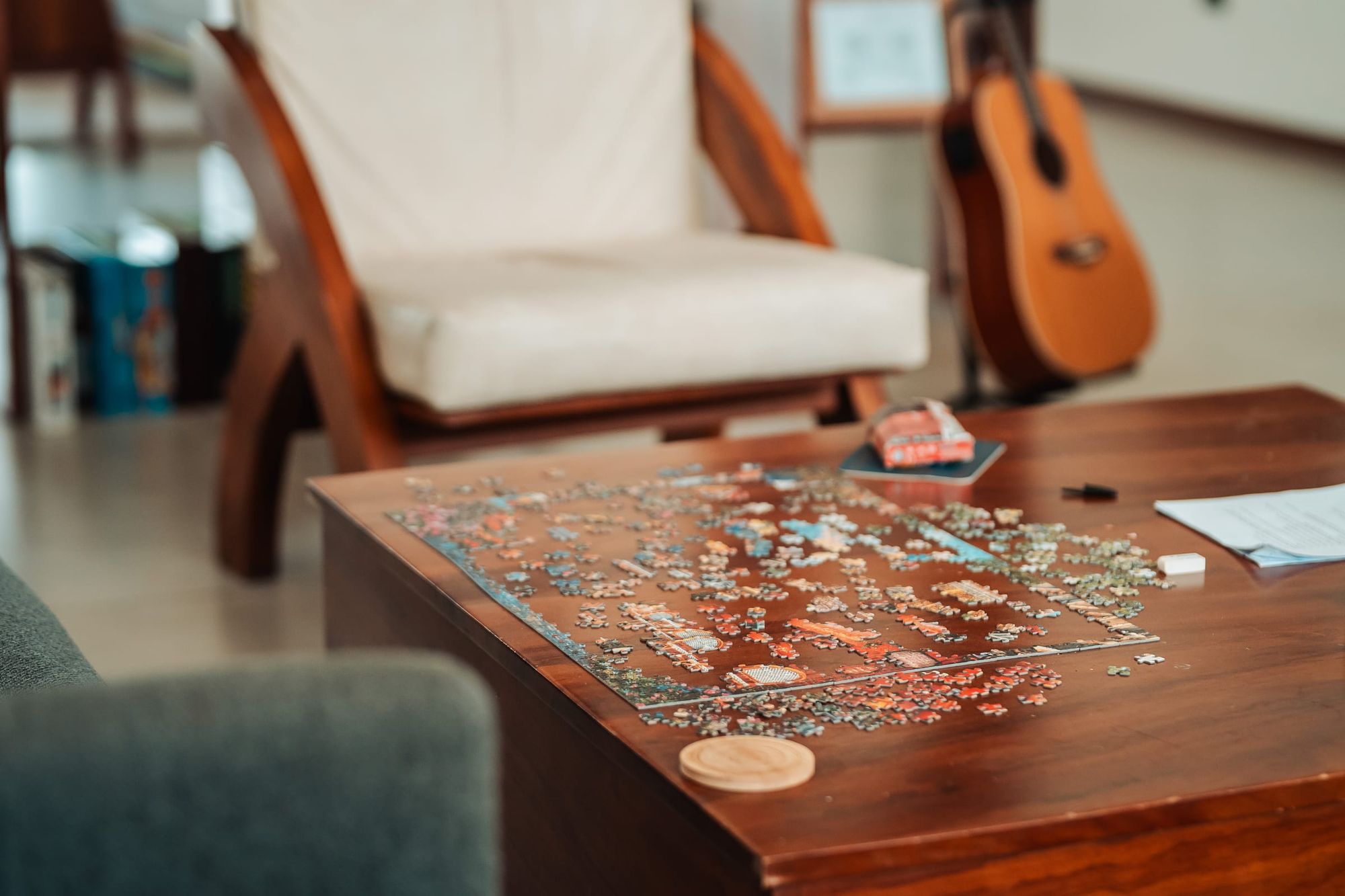 A half-finished jigsaw puzzle on a wooden table, with an armchair and a guitar behind.
