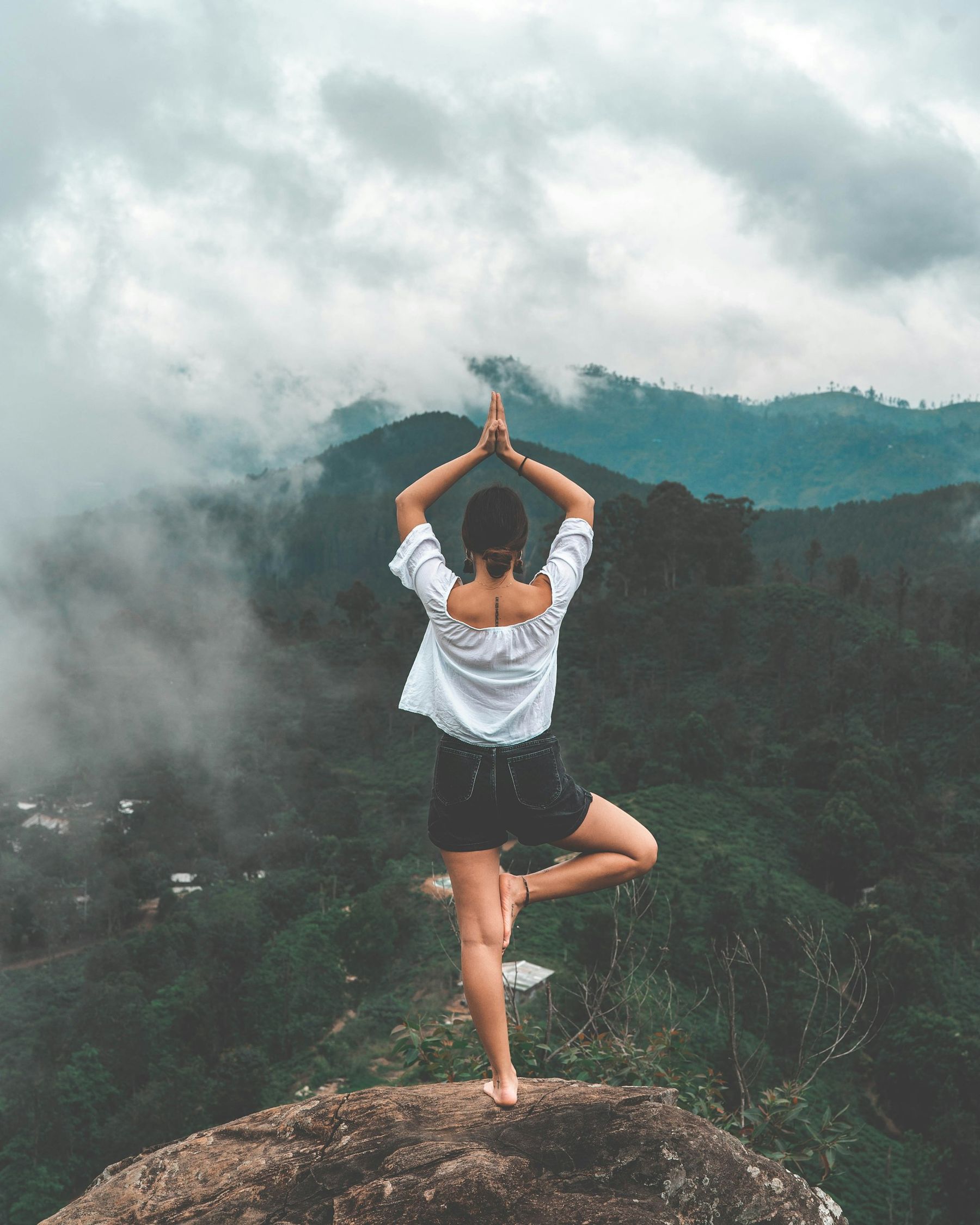 A person stands in a yoga tree pose on a rocky ledge, arms raised overhead, facing misty green mountains and forested hills.