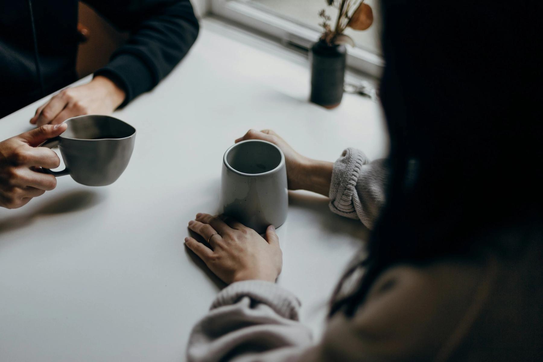 Two people sitting across a table in quiet conversation over warm mugs.