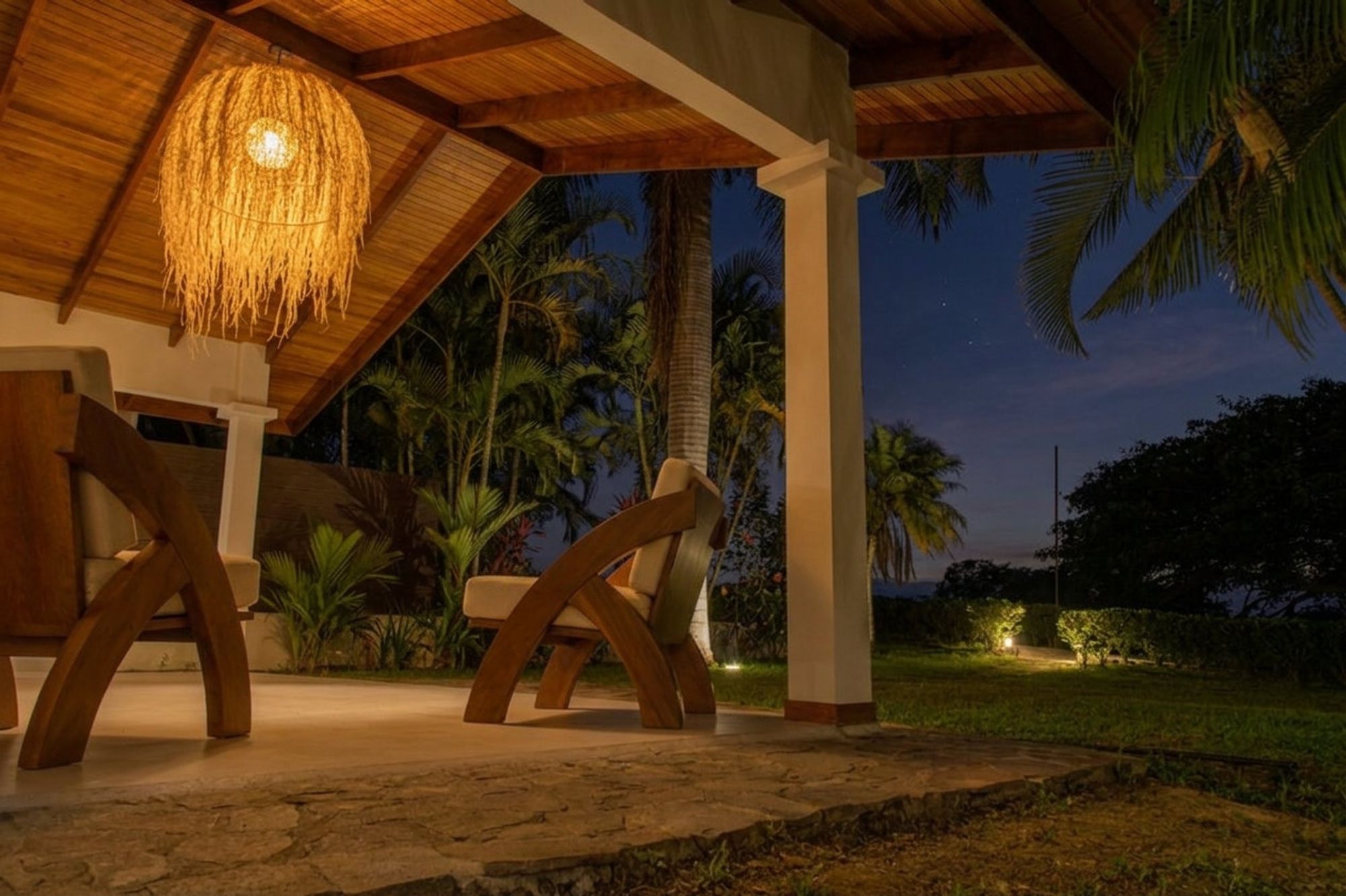 A covered terrace at dusk. A woven pendant lamp glows over two wooden lounge chairs, with palms and the valley beyond under a deep blue sky.
