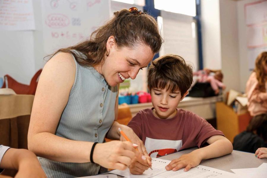 A Senesh teacher smiling and helping a kindergartener write.