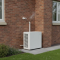 A modern white air source heat pump outdoor unit installed beside a UK semi detached brick home with a small gas boiler flue visible above on the wall, illustrating a hybrid heat pump configuration in 2026.