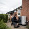 A qualified heating engineer servicing an air source heat pump outside a UK home while a homeowner watches from the doorway.