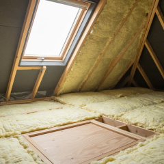 A clean UK loft interior in 2026 with thick mineral wool quilt laid in two crossed layers between and across the timber joists, soft natural light, a clear ventilation gap at the eaves, exposed roof rafters and felt, neat boarding around the loft hatch, illustrating a professional loft insulation install to current Building Regulations.