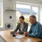 A bright modern UK home in 2026 being assessed for Warm Homes Local Grant upgrades, with an energy adviser reviewing documents beside visible loft insulation, solar panels, smart heating controls and an air source heat pump outside.