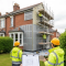 A UK terraced house being retrofitted under PAS 2035 with workers in hi vis jackets installing external wall insulation and a clipboard of technical drawings in the foreground.